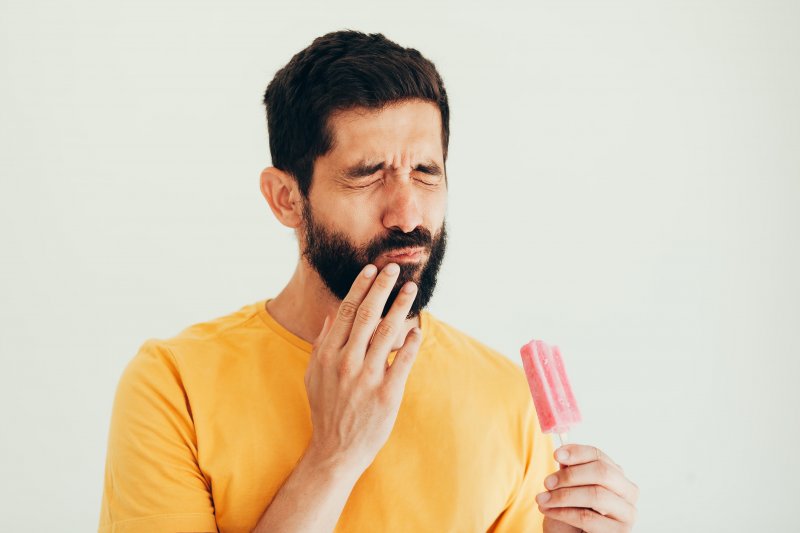 Bearded man in yellow shirt putting his hand up to his mouth and grimacing while holding a red popsicle